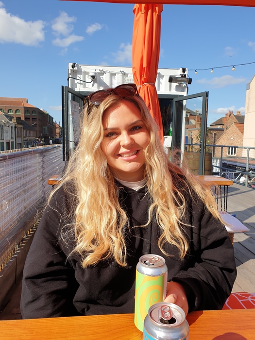 Young blonde white woman sat at an outdoor bar with a can of beer.