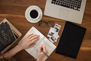 person writing in a journal with coffee and a laptop on a desk