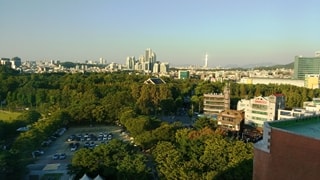 View from a rooftop over looking car park, buildings and large number of trees with the city of Seoul in the background