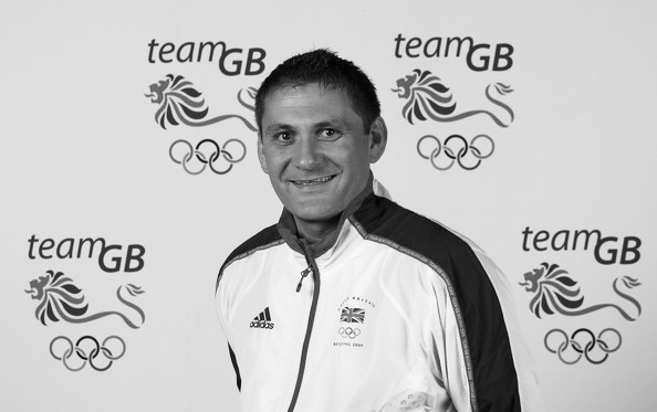 Adrian Hinchcliffe smiling in front of a background with the team GB logo on it for an Olympic games
